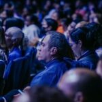 A large, diverse audience attentively listens during an indoor event.