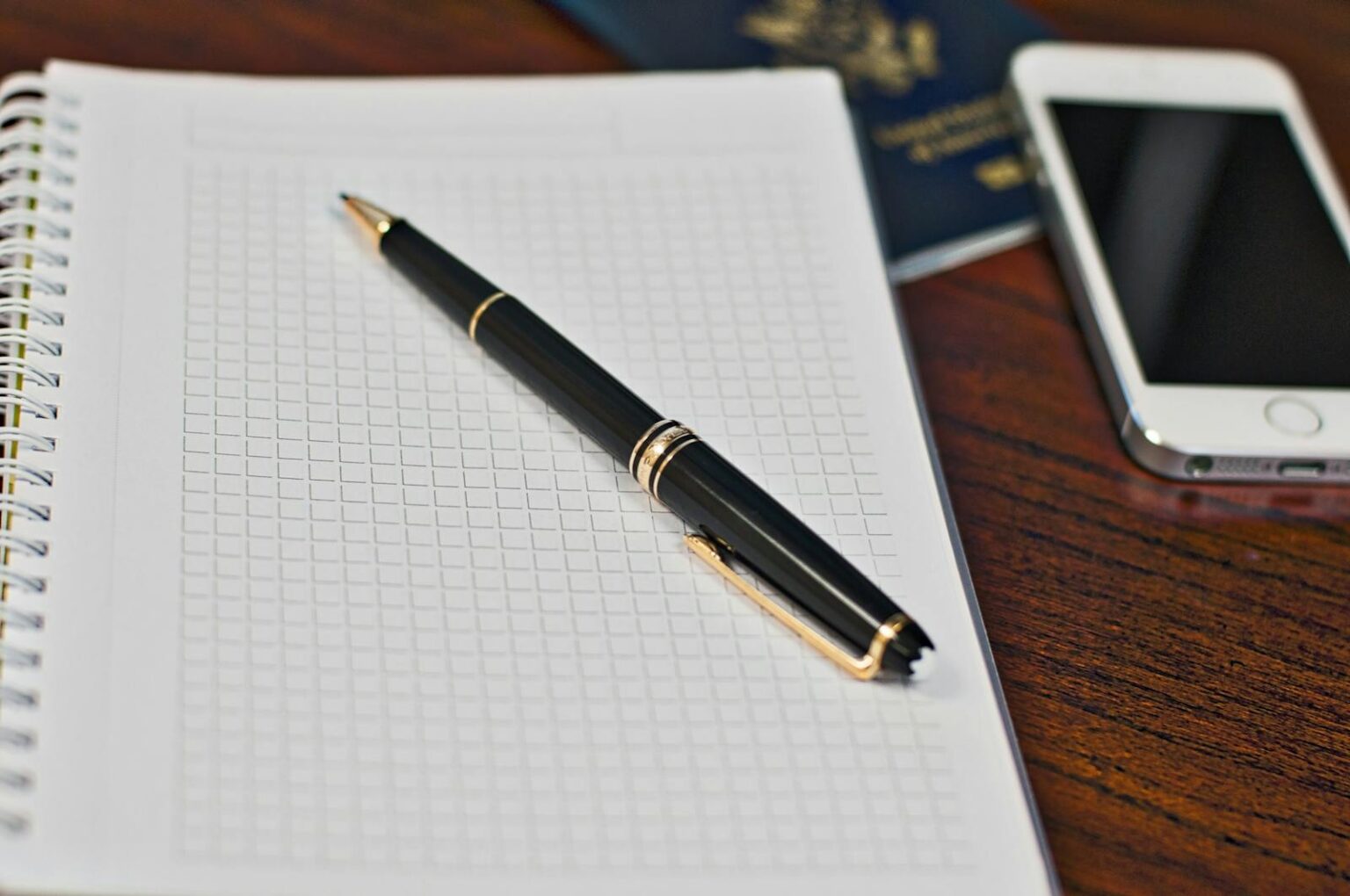 Black pen on a grid notebook with smartphone and passport on wooden desk.