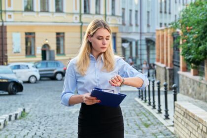 Młoda biznesowa kobieta w mieście patrząc na jej zegarek na rękę. Pewna siebie, poważna kobieta z papierami w dłoniach idąca ulicą