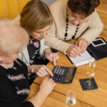 Widok z góry inteligentnych, ładnie wyglądających starszych kobiet spotykających się w nowoczesnej kawiarni z pomarańczowymi ścianami, mających na stole kalkulator i notatnik, omawiających ceny, liczenie i zapisywanie liczb