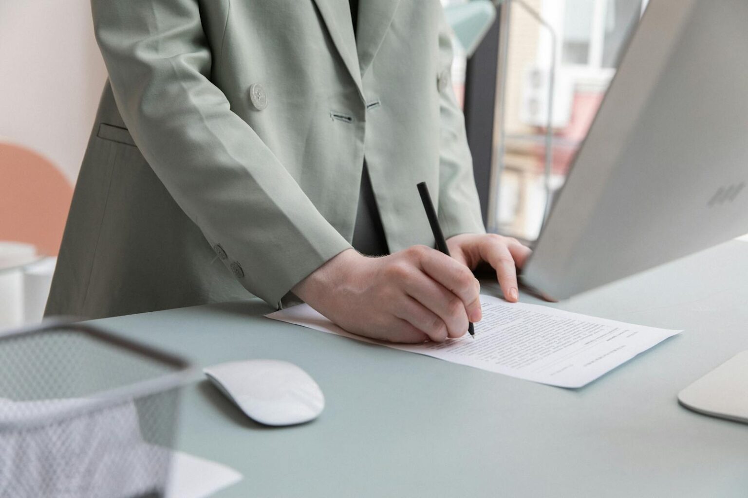 Unrecognizable worker standing at table with computer while taking notes in document while working in office